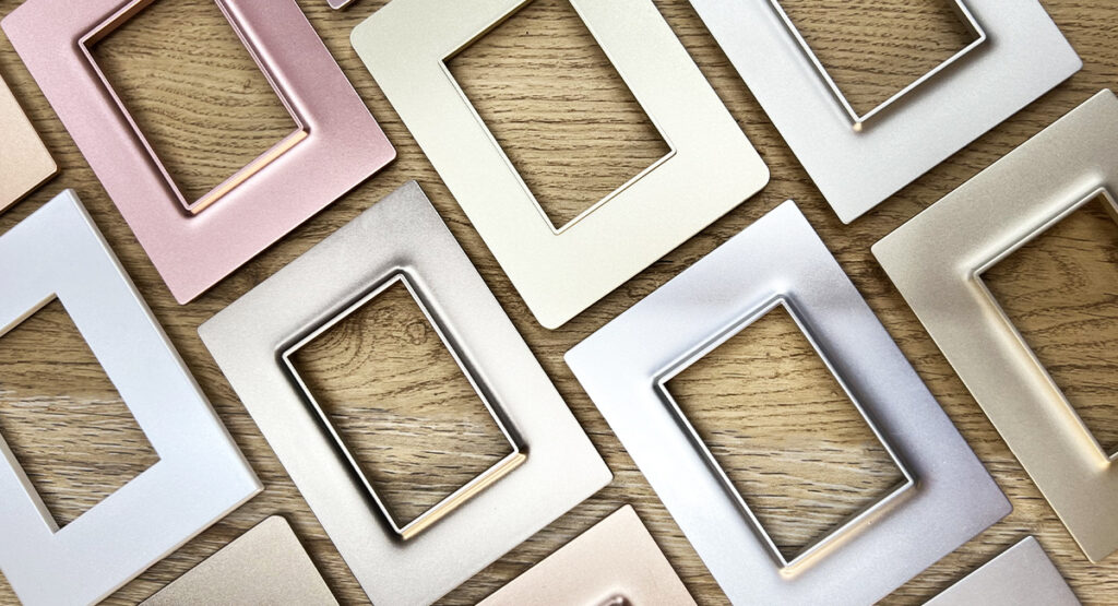 An assortment of square electrical switch plates in various metallic finishes, including rose gold, silver, and gold, arranged on a wooden surface.