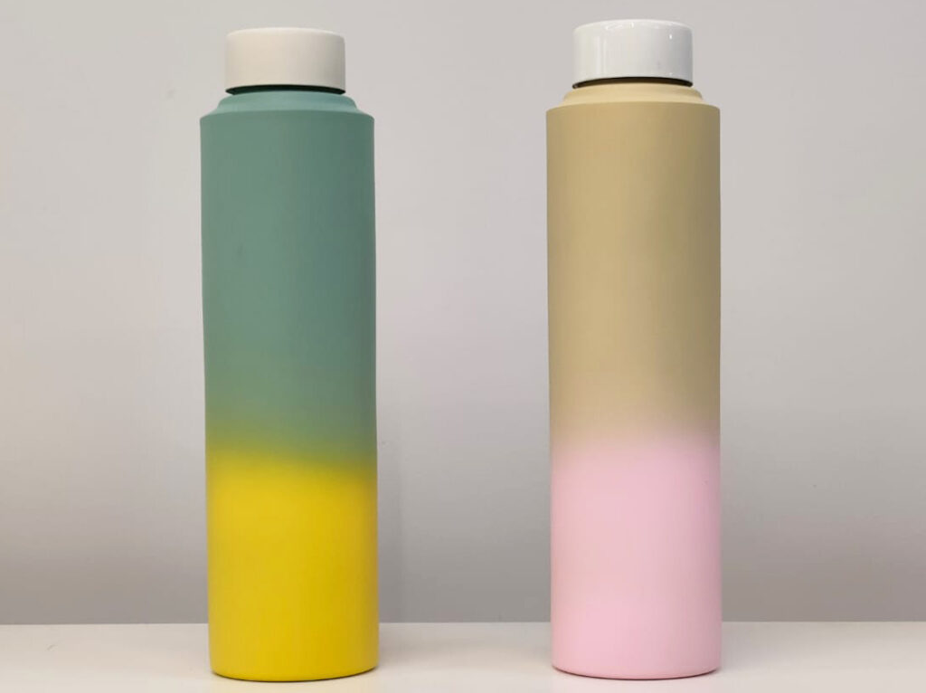 Two stainless steel water bottles with an ombre paint job, one transitioning from green to yellow and the other from beige to pink.