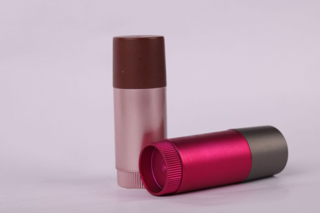 Two cylindrical cosmetic containers with spray-painted lids in metallic pink and brown, and bases in metallic gray and pink.