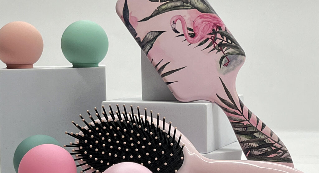 A pink hairbrush with a flamingo and tropical leaf pattern, a smaller hairbrush with gold bristles, and two green and pink hair rollers.